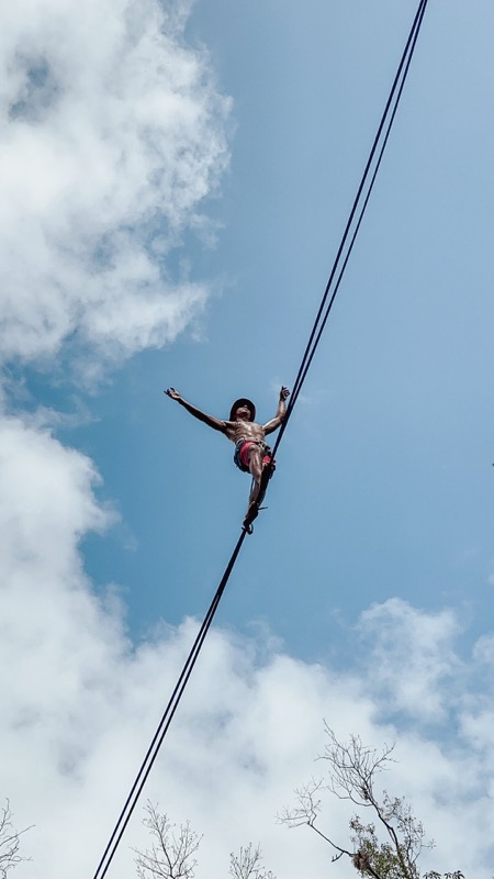 A slackliner walks a highline against a blue sky and clouds, arms out for balance.