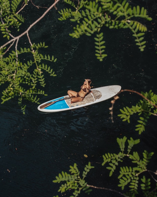 From above, a woman lies on a paddleboard on the dark water of a cenote, framed by leaves.