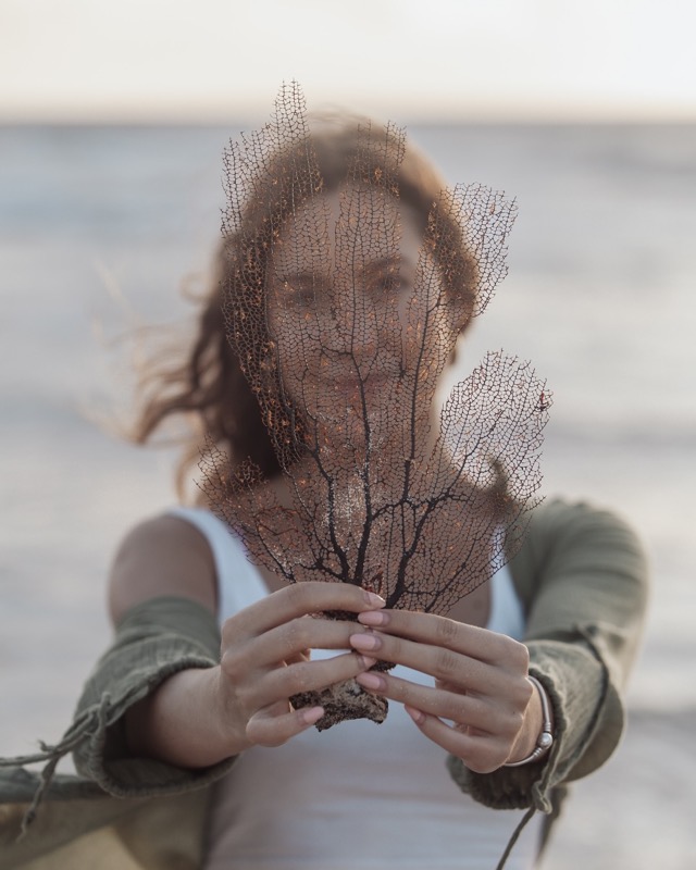 A woman holds a dried sea fan in front of her face on the beach.