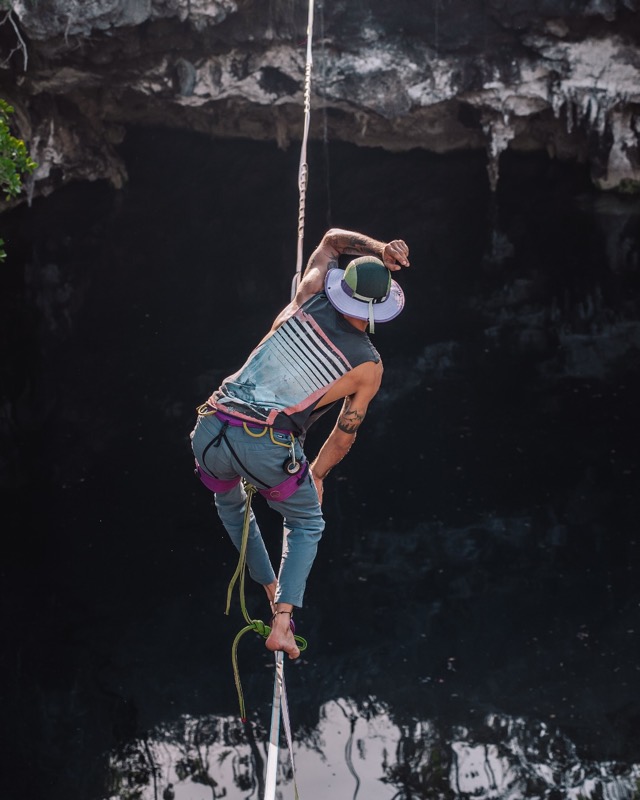 A slackliner in a harness balances on a line strung over a dark cenote.