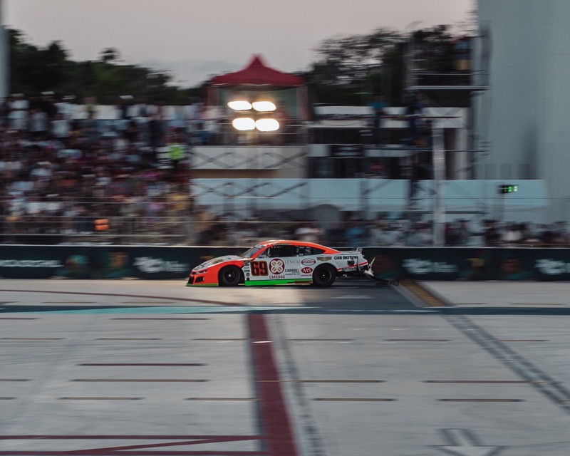 A race car speeds past packed grandstands at dusk on a runway turned racetrack in Tulum.